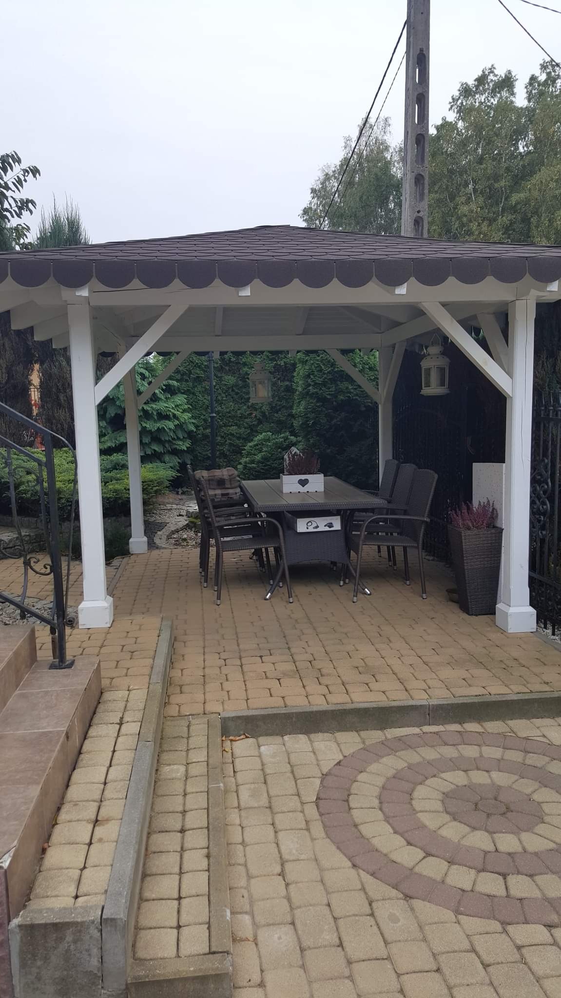 Biała altana z brązowym dachem, stół z krzesłami na kostce brukowej, dekoracje na stole i wisząca latarenka.