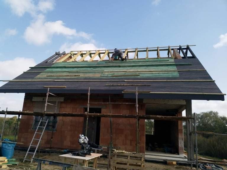 Budowa dachu: dekarz pracuje na częściowo pokrytym dachu budynku z czerwonej cegły, widoczne rusztowania, narzędzia na stole i niebieskie niebo z chmurami.