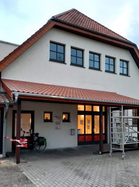 Zadaszone wejście do budynku z czerwonymi drzwiami i rusztowaniem po prawej stronie, rower zaparkowany po lewej, całość oświetlona w pochmurny dzień.