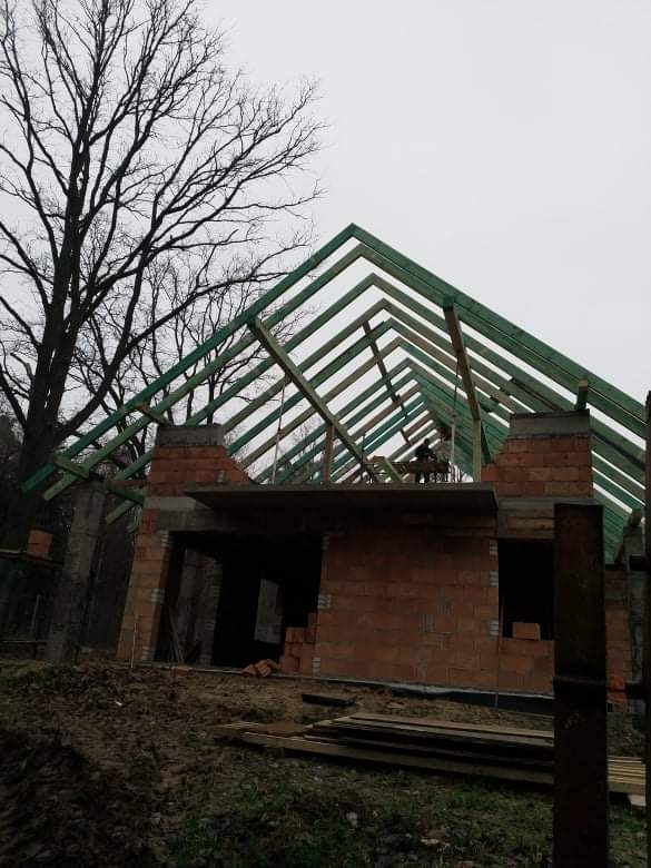 Budowa domu jednorodzinnego w stanie surowym otwartym: widoczne czerwone cegły, drewniana konstrukcja dachu w kolorze zielonym, w tle nagie drzewa.