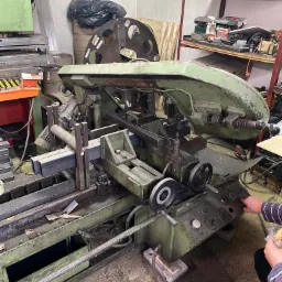 Automatyczna piła taśmowa do metalu w warsztacie, z zamocowanym profilem stalowym i dłonią operatora naciskającą przycisk sterujący.