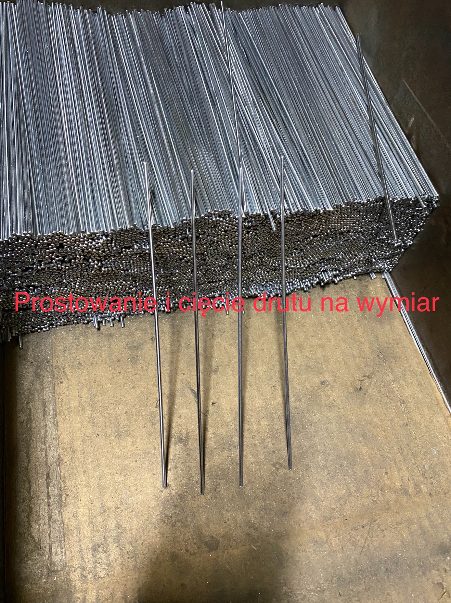 Stos srebrnych, prostych prętów metalowych, ułożonych równolegle na szorstkiej, brązowej powierzchni. Kilka prętów wystaje pionowo ponad resztę.