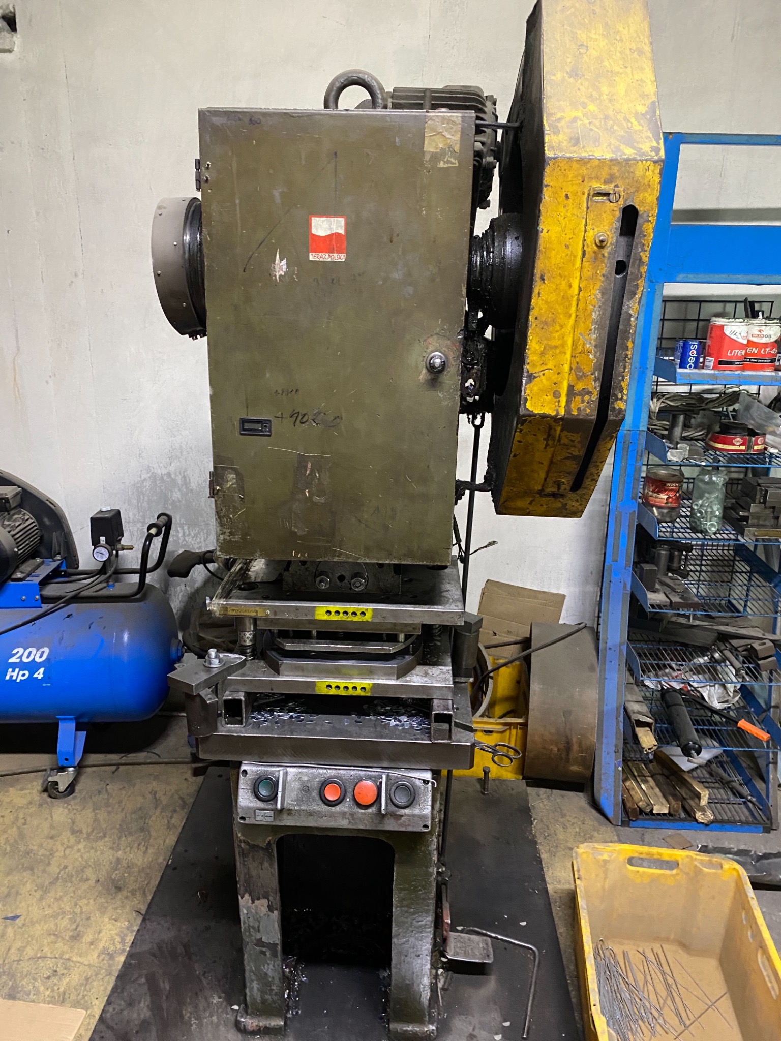 Przemysłowa prasa mechaniczna w warsztacie, widoczne elementy sterujące, osłona z żółtą farbą, obok niebieski kompresor i regał z narzędziami.