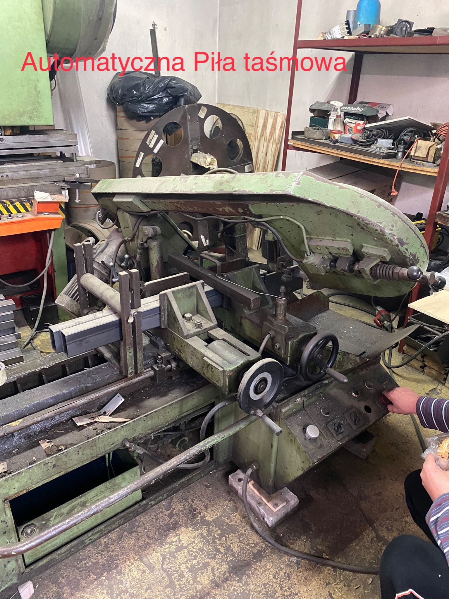 Automatyczna piła taśmowa do metalu w warsztacie, z zamocowanym profilem stalowym i dłonią operatora naciskającą przycisk sterujący.