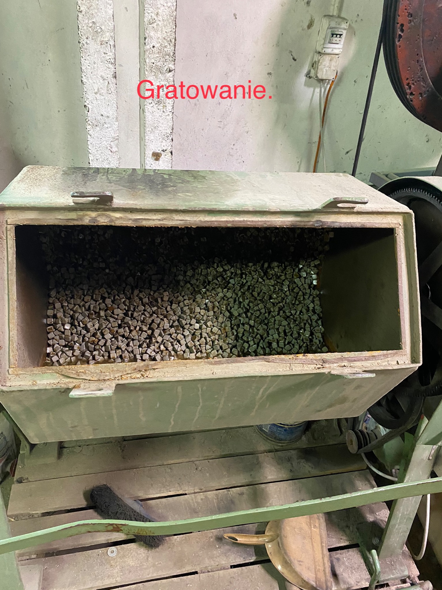 Otwarty pojemnik maszyny do gratowania wypełniony drobnymi, szarymi elementami ściernymi, z widocznym fragmentem silnika i instalacji elektrycznej w tle.