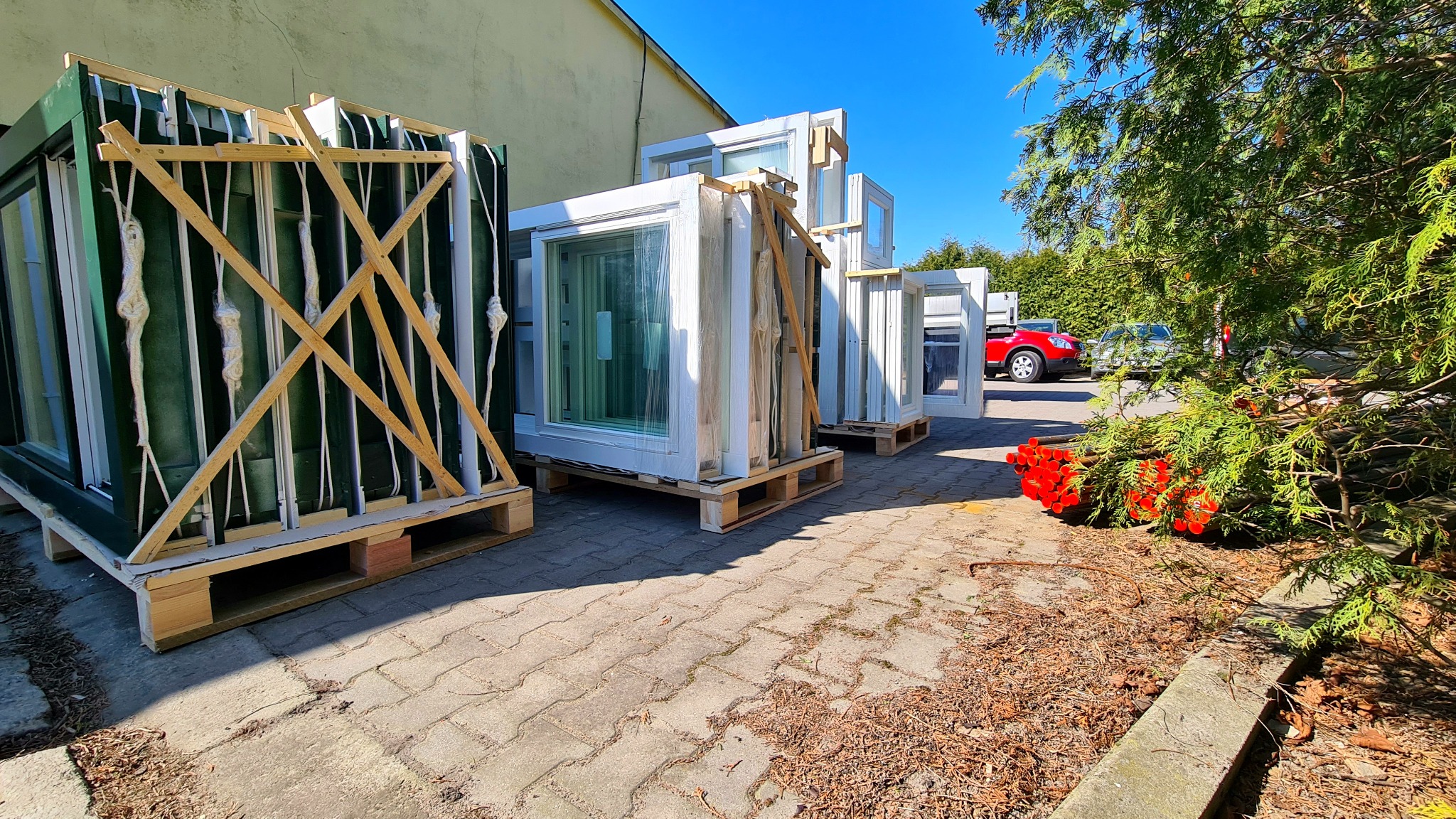 Sztaple okien na paletach, zabezpieczone drewnianymi listwami i folią, czekają na montaż na zewnątrz budynku w słoneczny dzień.