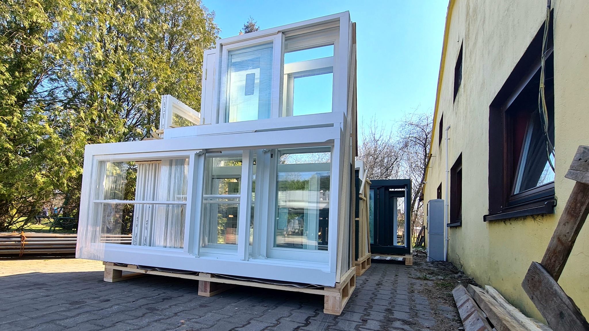 Białe ramy okienne i drzwiowe ułożone na paletach przed budynkiem, niektóre owinięte folią zabezpieczającą, w tle drzewa i błękitne niebo.