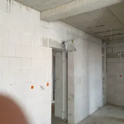 Wnętrze w stanie surowym z widoczną instalacją elektryczną: kable prowadzone po ścianach z betonu komórkowego, puszki elektryczne w kolorze pomarańczowym, wiązka kabli nad otworem drzwiowym.