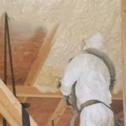 Pracownik w kombinezonie ochronnym aplikuje pianę izolacyjną na poddaszu, widoczne drewniane krokwie i pokryta pianą powierzchnia dachu.