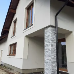 Wykończona elewacja domu jednorodzinnego z jasnoszarym tynkiem, drewnianymi oknami i kamienną okładziną na filarze wejściowym, widoczne rury spustowe i przygotowany teren wokół budynku.