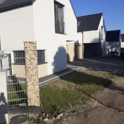 Nowoczesne domy jednorodzinne z białymi elewacjami i grafitowymi dachami, widoczne ogrodzenie z gabionami i elementami kamiennymi, niezagospodarowany teren wokół.