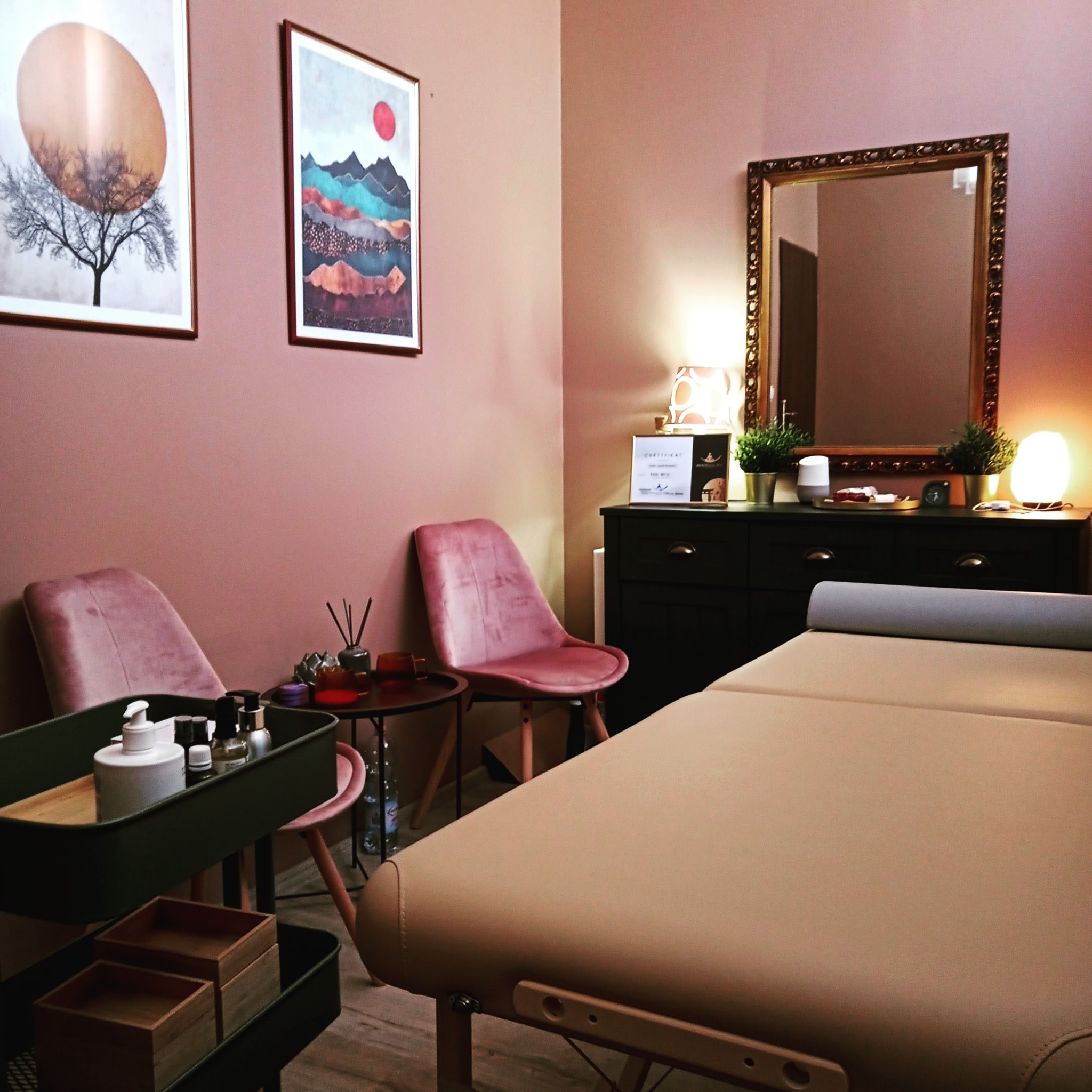 Gabinet masażu z beżowym łóżkiem do masażu, różowymi krzesłami, czarną komodą z lustrem w złotej ramie i dekoracjami, na ścianach wiszą obrazy z motywami drzew i gór.