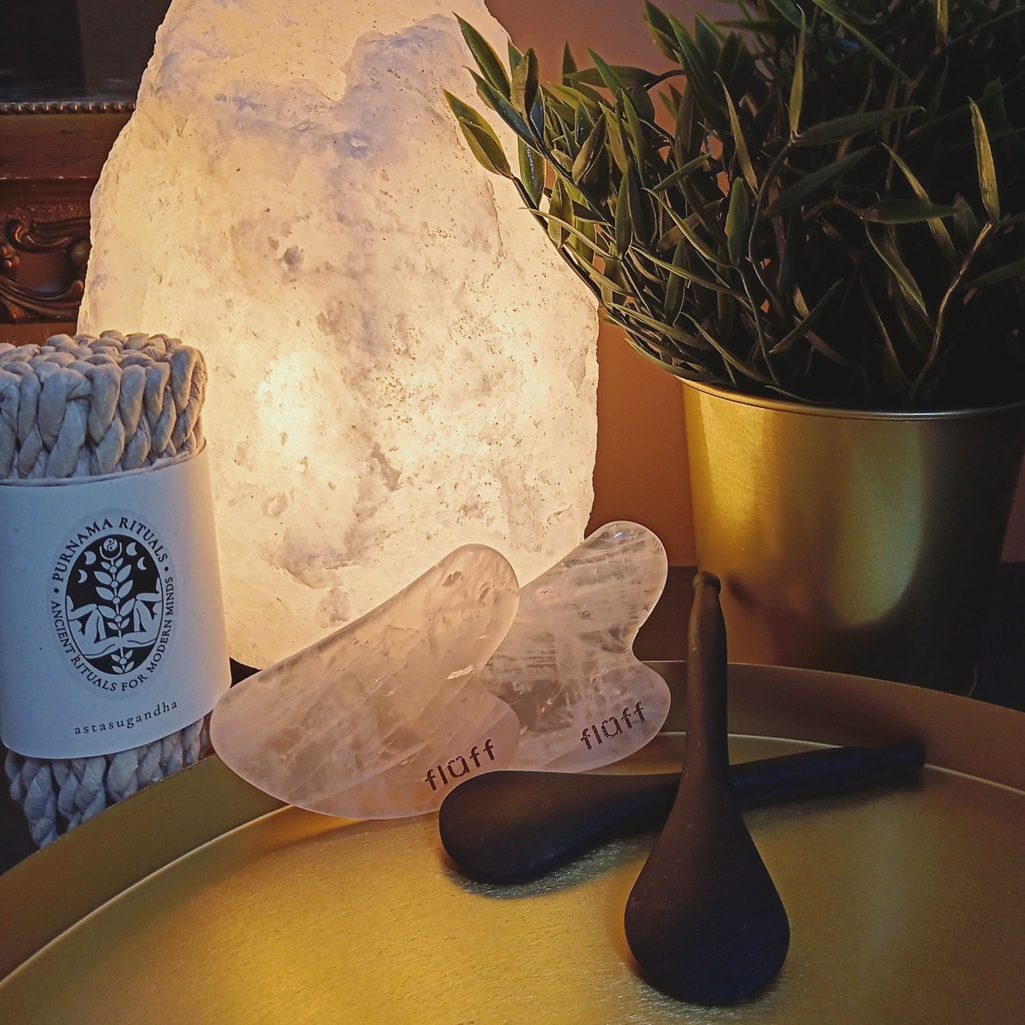 Zestaw do masażu twarzy: dwa kamienie gua sha z kwarcu różowego marki Fluff, czarne kamienne łyżeczki do nakładania kosmetyków, obok zapalona lampa solna i doniczka z rośliną na złotej tacy.