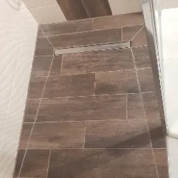 Nowoczesna kabina prysznicowa z odpływem liniowym w podłodze, wykończona brązowymi płytkami imitującymi drewno, kontrastującymi z białymi płytkami na ścianach.