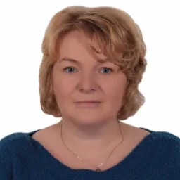 Portret kobiety o blond włosach i niebieskich oczach, ubrana w niebieski sweter, z delikatnym srebrnym łańcuszkiem na szyi, na białym tle.