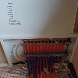 Rozdzielacz ogrzewania podłogowego w szafce z listą pomieszczeń wypisaną ręcznie na drzwiczkach, widoczne rury doprowadzające wodę do poszczególnych pętli grzewczych, folia izolacyjna z nadrukowaną...