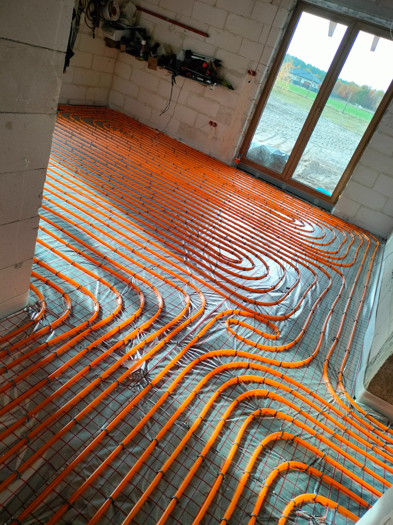 Pomarańczowe rury ogrzewania podłogowego ułożone spiralnie na folii izolacyjnej w surowym wnętrzu z widokiem na zewnątrz przez okno.