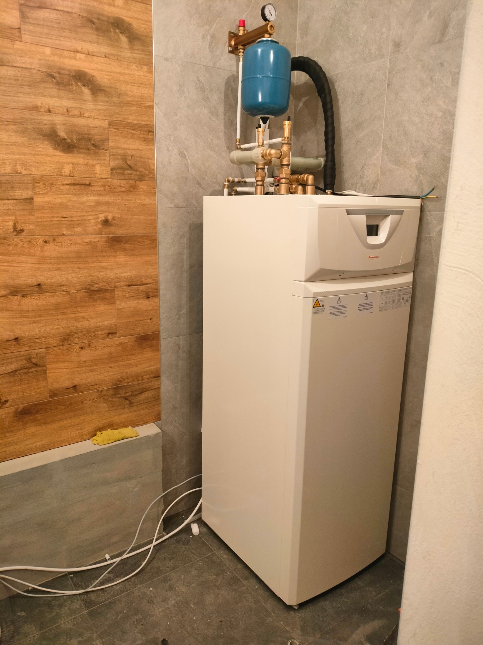 Biały, nowoczesny piec gazowy z niebieskim zbiornikiem wyrównawczym i miedzianymi rurami, zamontowany na tle szarej płytki i drewnopodobnej okładziny ściennej.