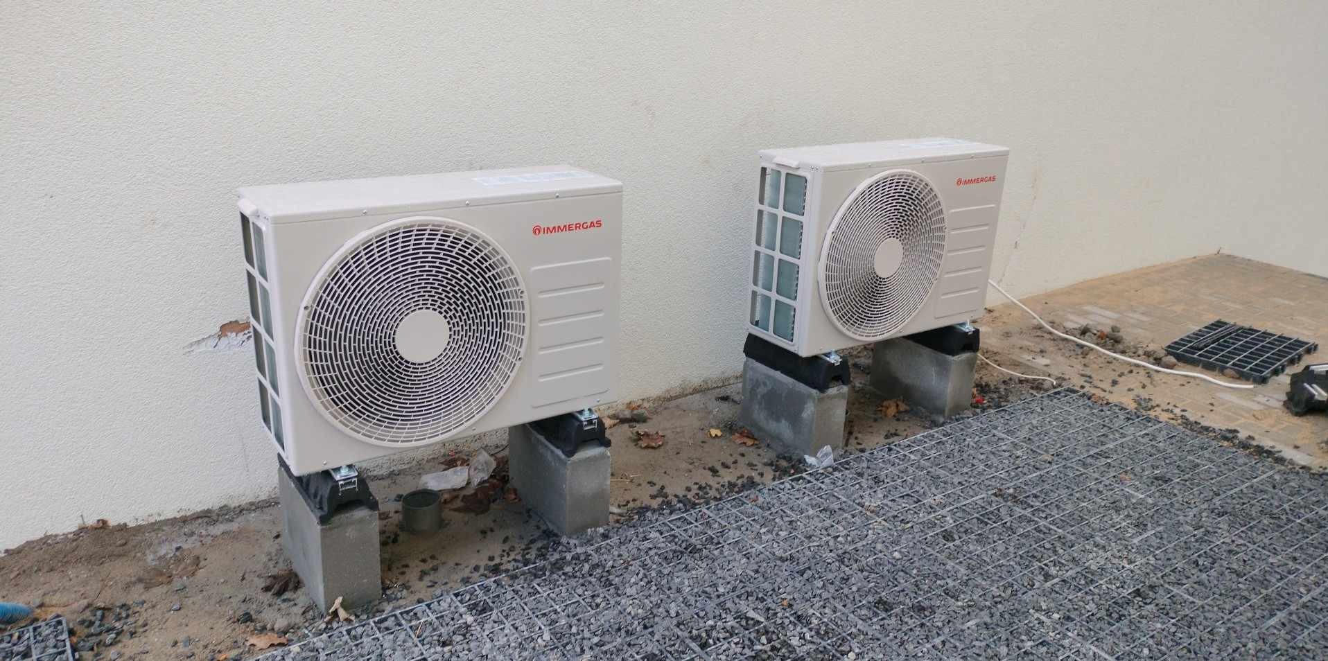 Dwa białe, zewnętrzne jednostki klimatyzacyjne marki Immergas, zamontowane na betonowych bloczkach, na żwirowym podłożu przy jasnej ścianie budynku.