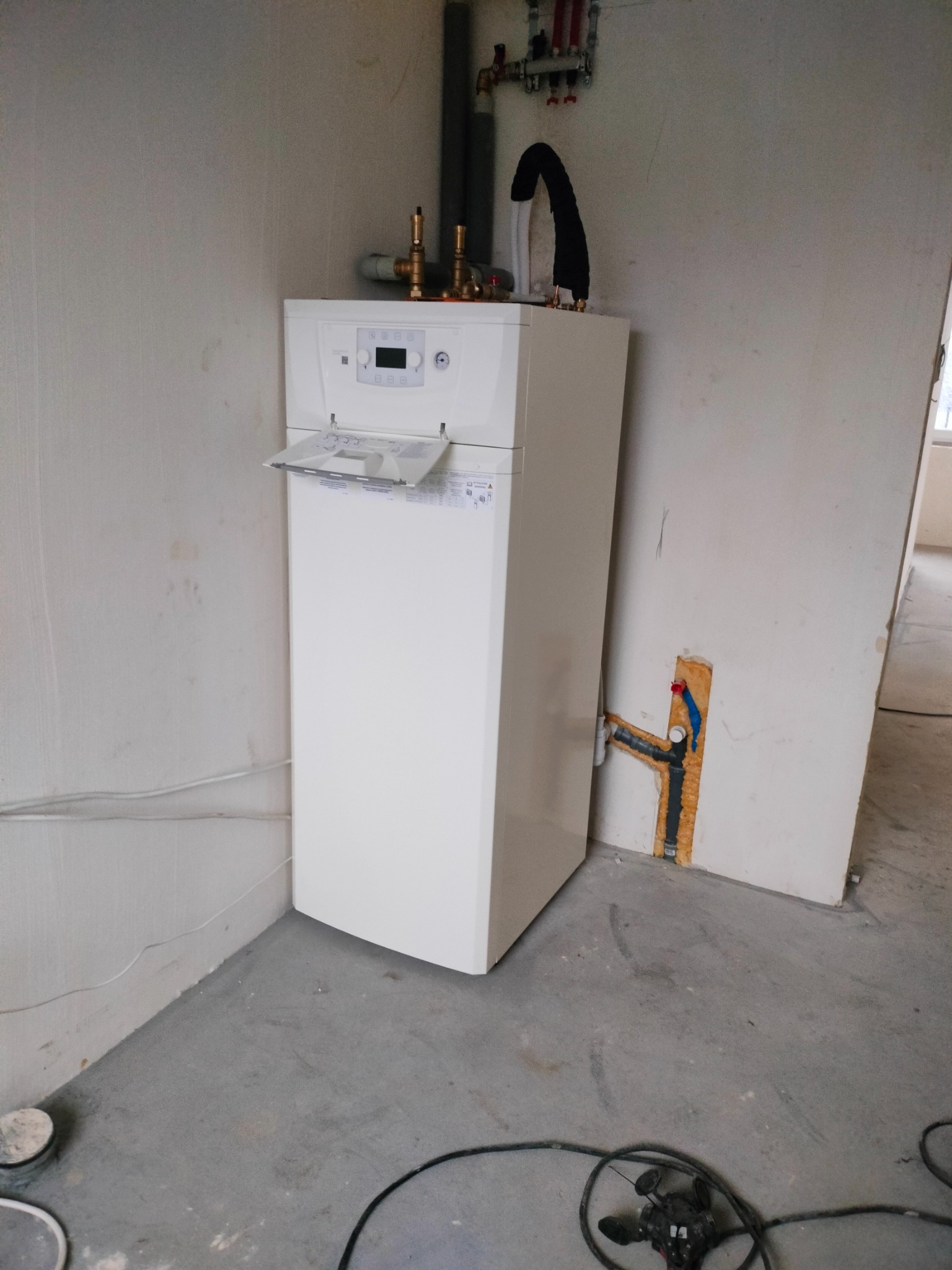 Nowy, biały piec gazowy z otwartą klapką, zainstalowany w rogu pomieszczenia w trakcie remontu, widoczne rury i instalacje na ścianie, surowy beton na podłodze.