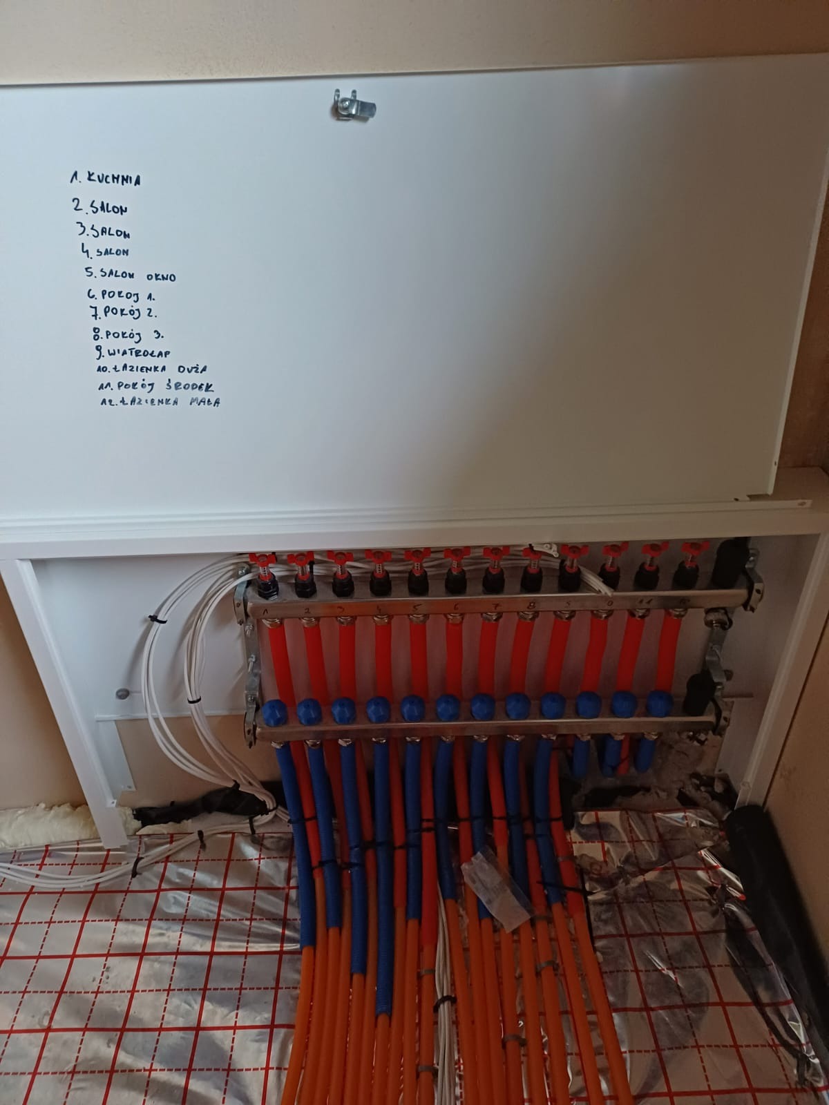 Rozdzielacz ogrzewania podłogowego w szafce z listą pomieszczeń wypisaną ręcznie na drzwiczkach, widoczne rury doprowadzające wodę do poszczególnych pętli grzewczych, folia izolacyjna z nadrukowaną...