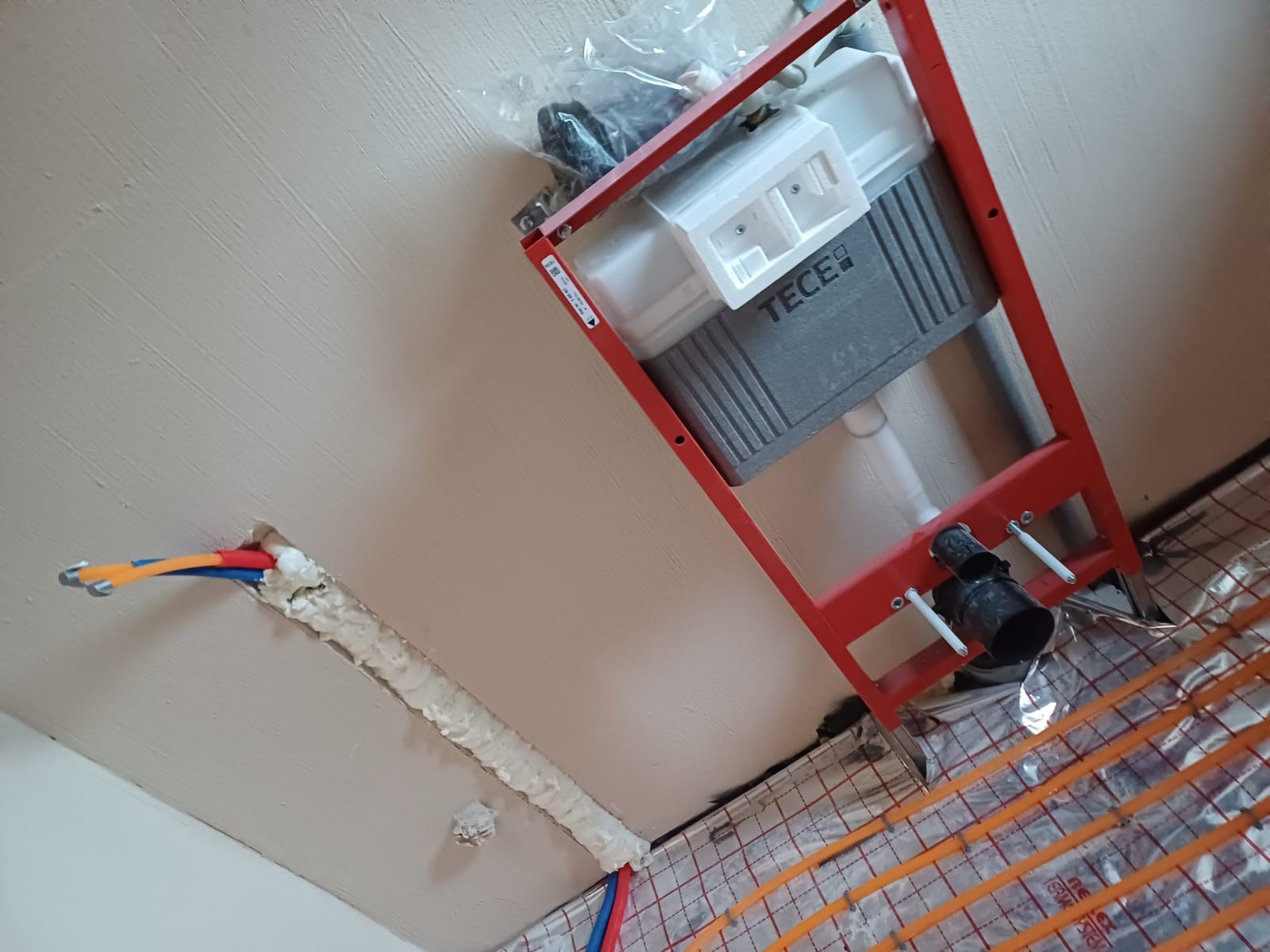 Stelaż podtynkowy TECE w czerwonej ramie, widoczne rury instalacyjne w ścianie zabezpieczone pianką montażową oraz ogrzewanie podłogowe w folii aluminiowej.