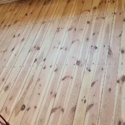 Ściana wyłożona jasnymi, pionowo ułożonymi deskami z widocznymi gwoździami, w dolnym lewym rogu widoczne narzędzia i zwinięty czerwony kabel.