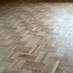 Ułożony parkiet z jasnego drewna w jodełkę francuską, widoczne niedoskonałości ściany w tle.