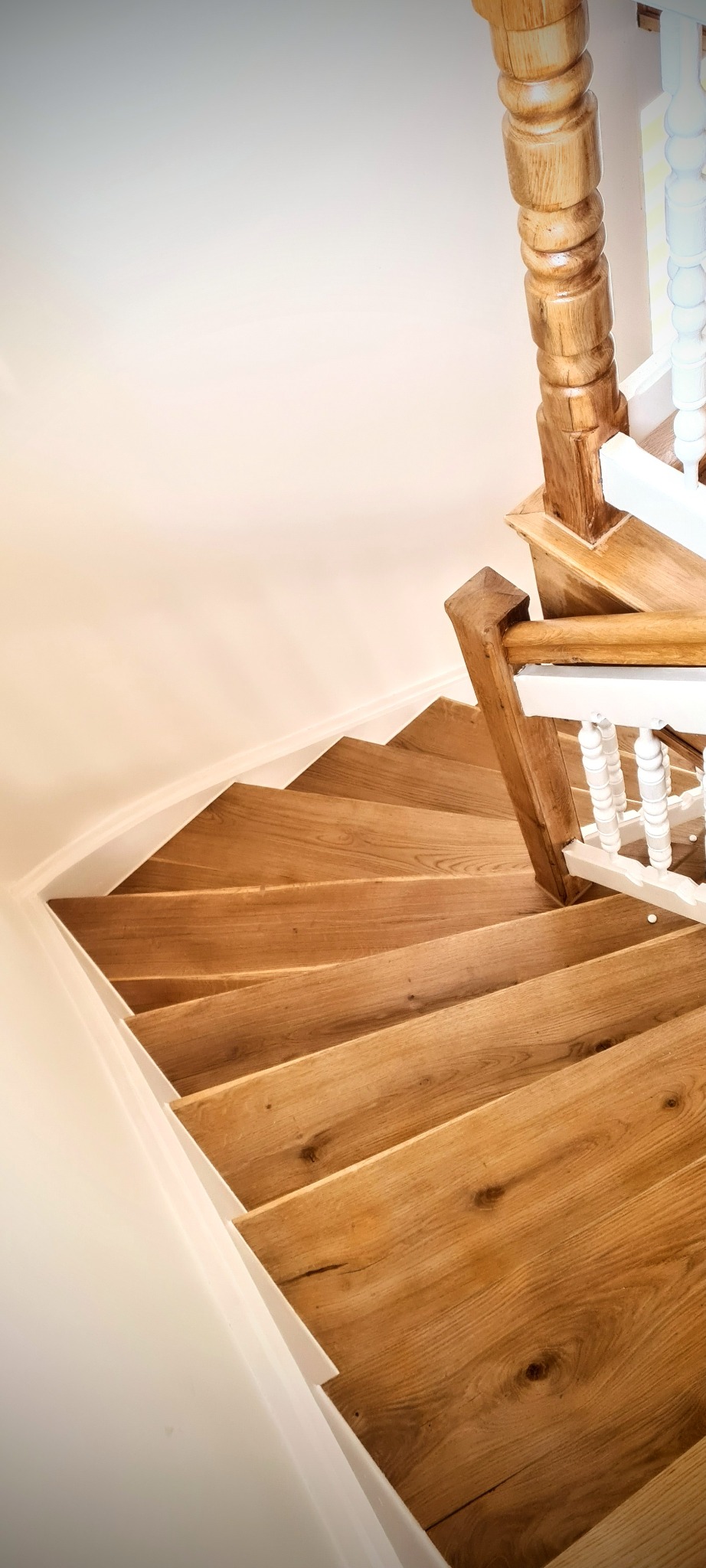 Widok z góry na kręte drewniane schody z jasnego drewna, z balustradą z drewna i białymi szczeblami, przylegające do zakrzywionej ściany w jasnym odcieniu.