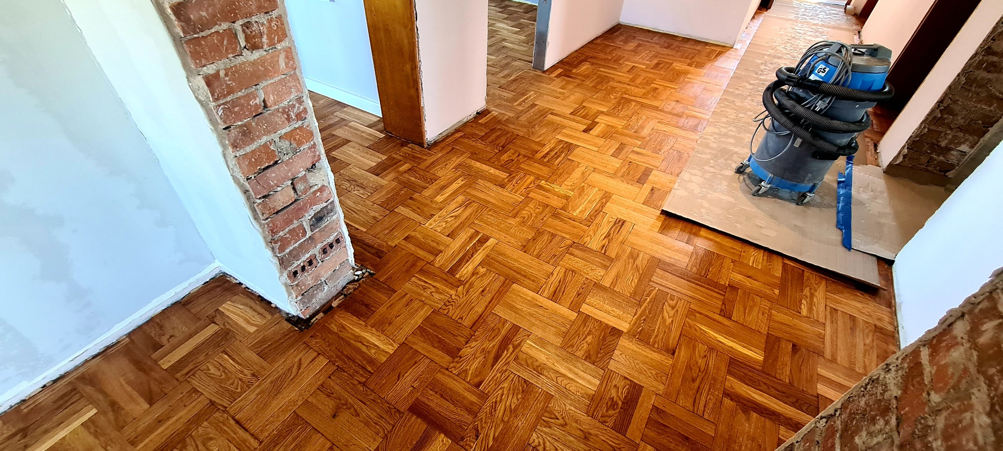 Parkiet ułożony w jodełkę, częściowo przykryty papierem ochronnym, z widocznym odkurzaczem przemysłowym oraz fragmentem ceglanego filaru w surowym stanie.