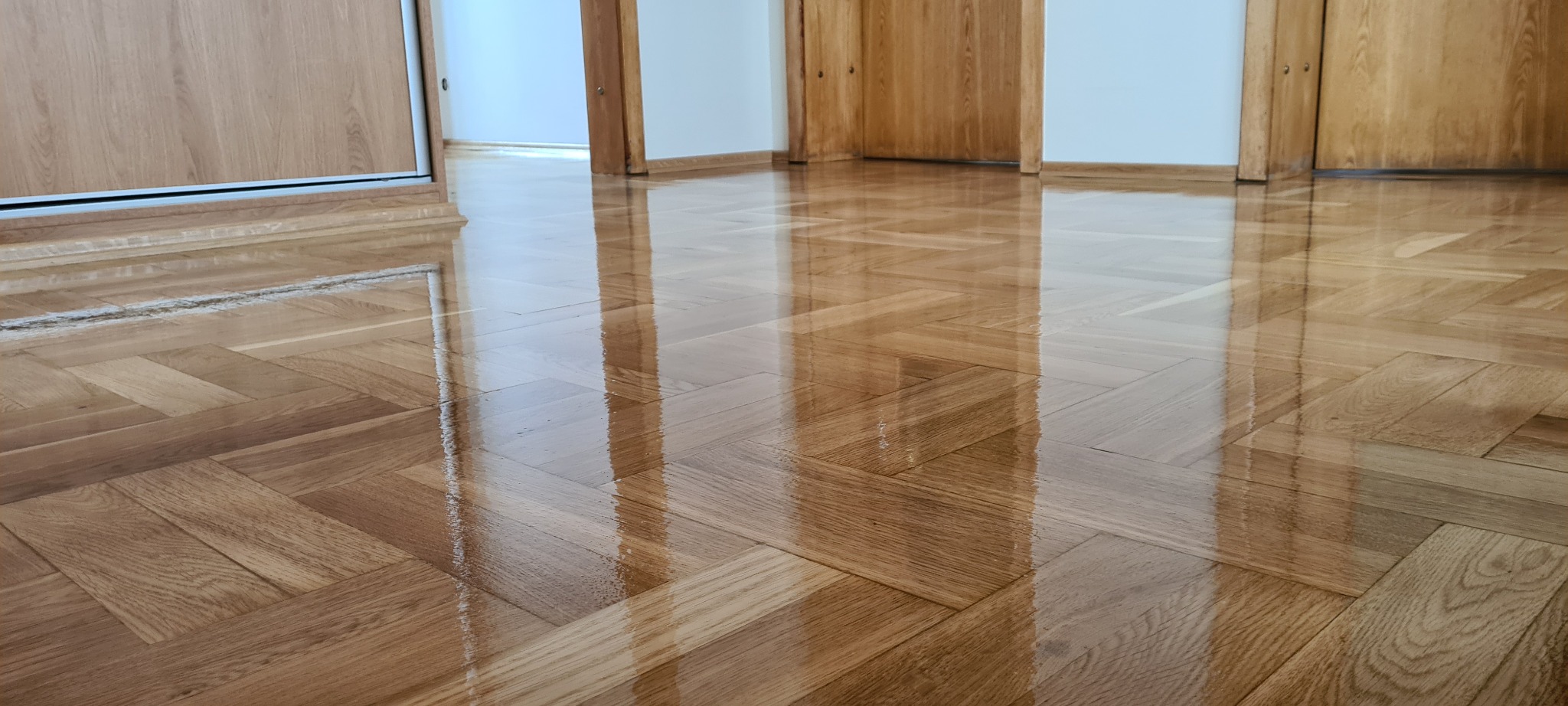 Świeżo polakierowany parkiet ułożony w jodełkę, odbijający światło i fragmenty drzwi w tle.