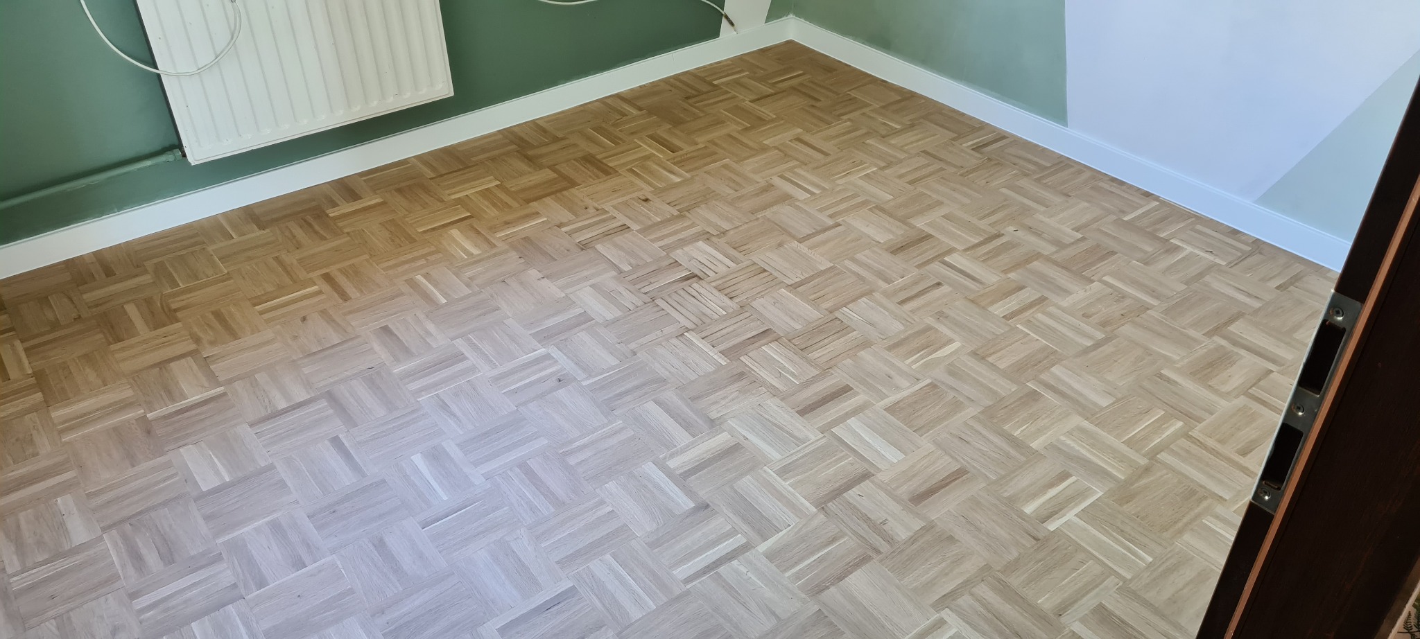 Jasny parkiet ułożony w kwadraty, widoczny fragment ściany pomalowanej na zielono i biała listwa przypodłogowa, fragment ościeżnicy drzwi.