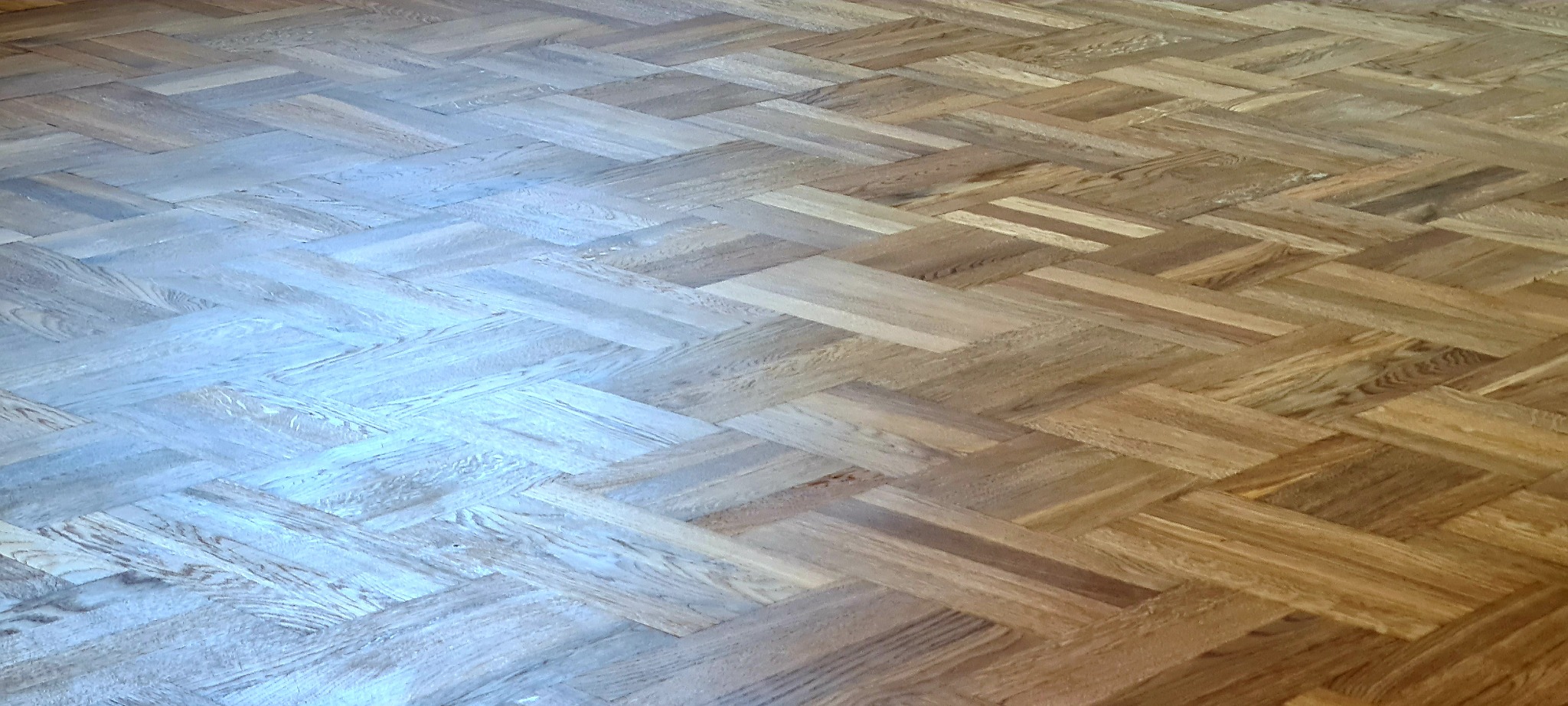 Parkiet ułożony w jodełkę, z widoczną różnicą w odcieniach po ewentualnym procesie renowacji lub czyszczenia, od jasnego, prawie bielonego drewna do naturalnego, ciepłego odcienia dębu.