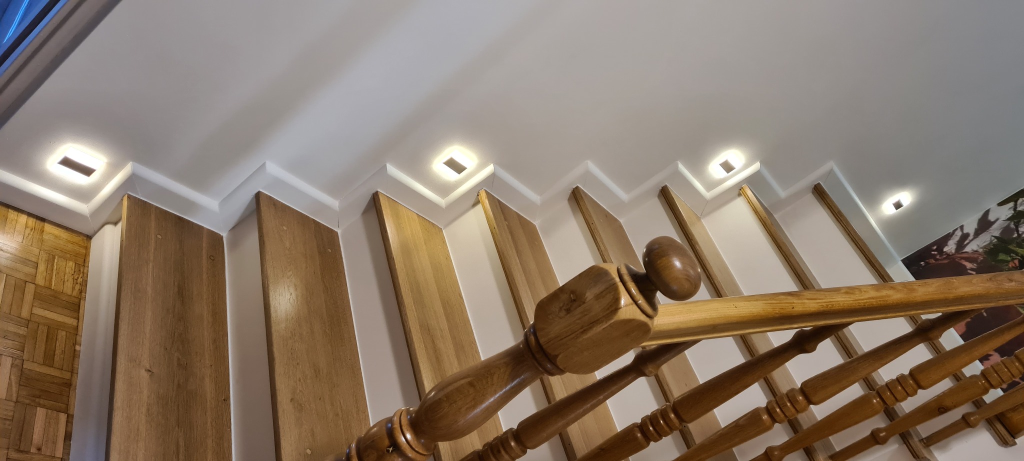 Drewniane schody z jasnymi stopniami i balustradą, oświetlenie punktowe w suficie, widok z dołu.