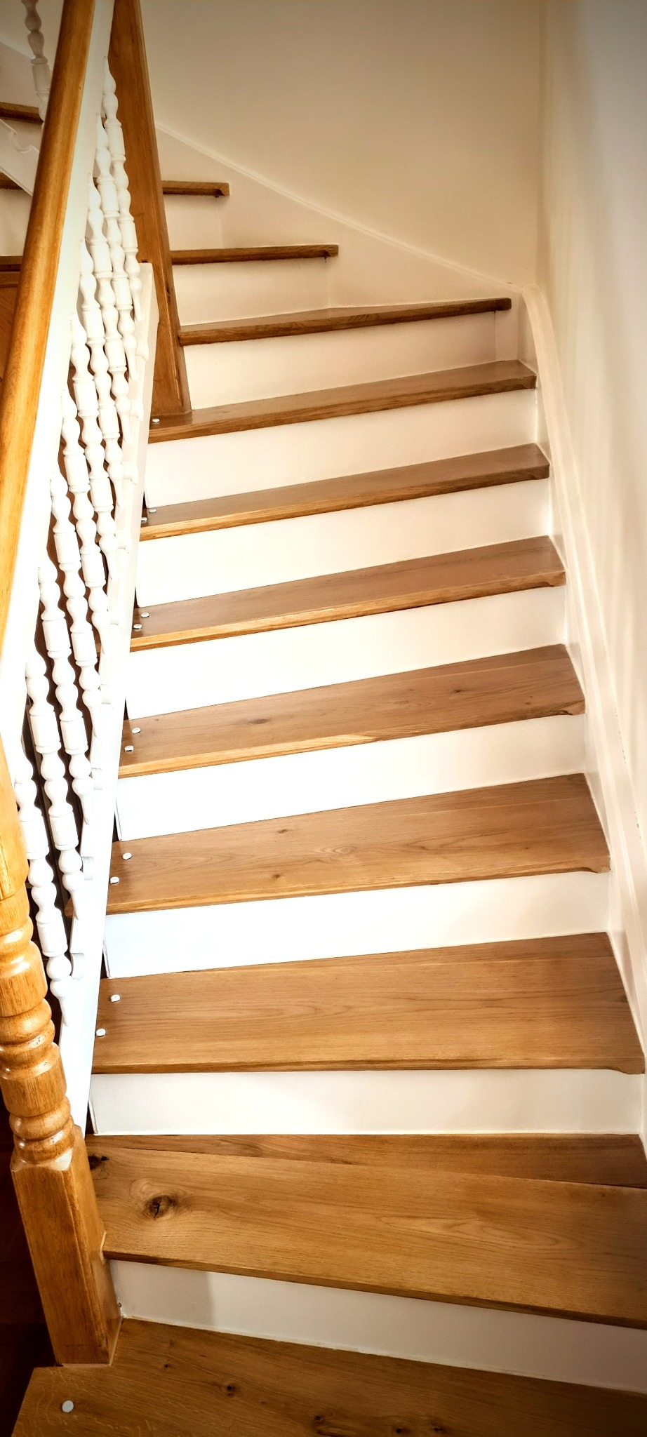 Bieg schodów z drewnianymi stopniami o jasnym odcieniu i białymi podstopnicami, balustrada z toczonymi elementami. Widoczne detale wykończenia.