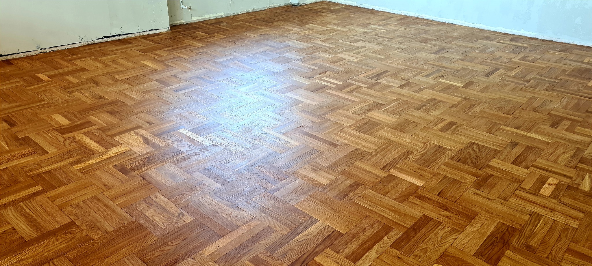 Ułożona parkietowa podłoga w jodełkę francuską, widoczne niedoskonałości ściany przy podłodze, jasne oświetlenie górne.