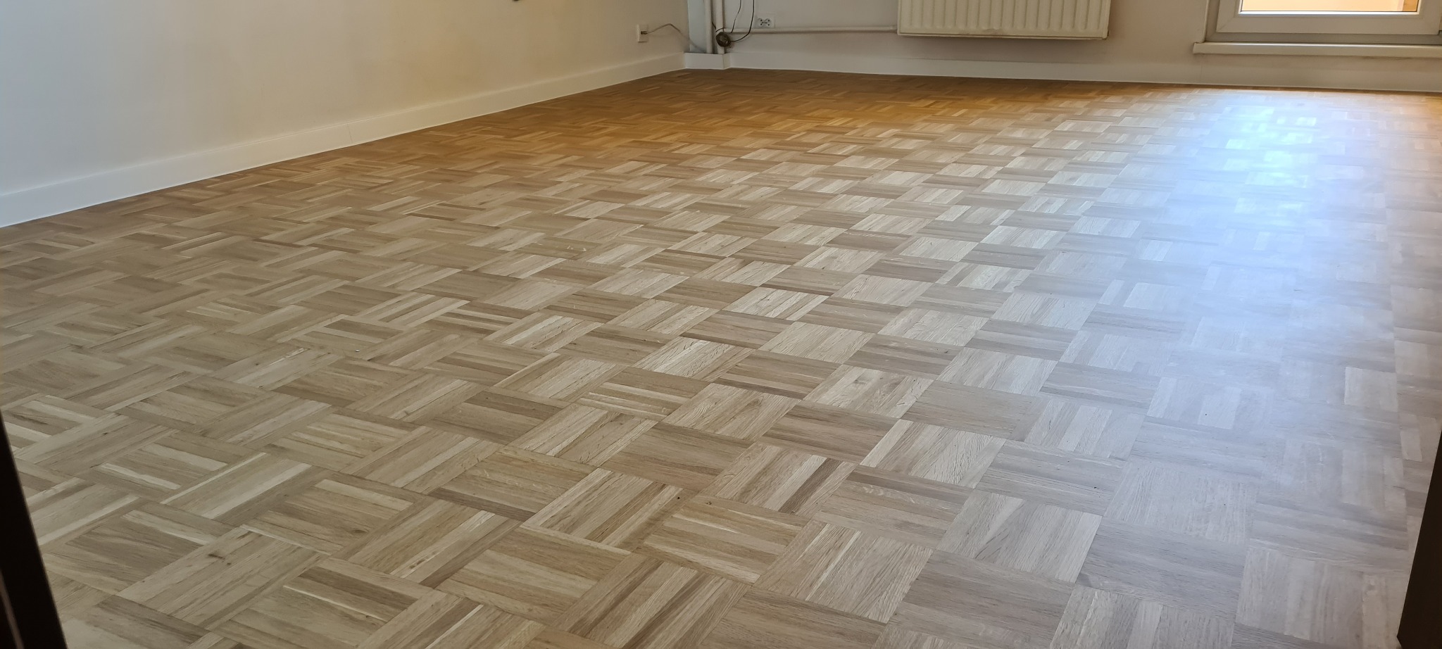 Parkiet ułożony we wzór szachownicy z jasnego drewna, widoczne listwy przypodłogowe i fragment okna z grzejnikiem.