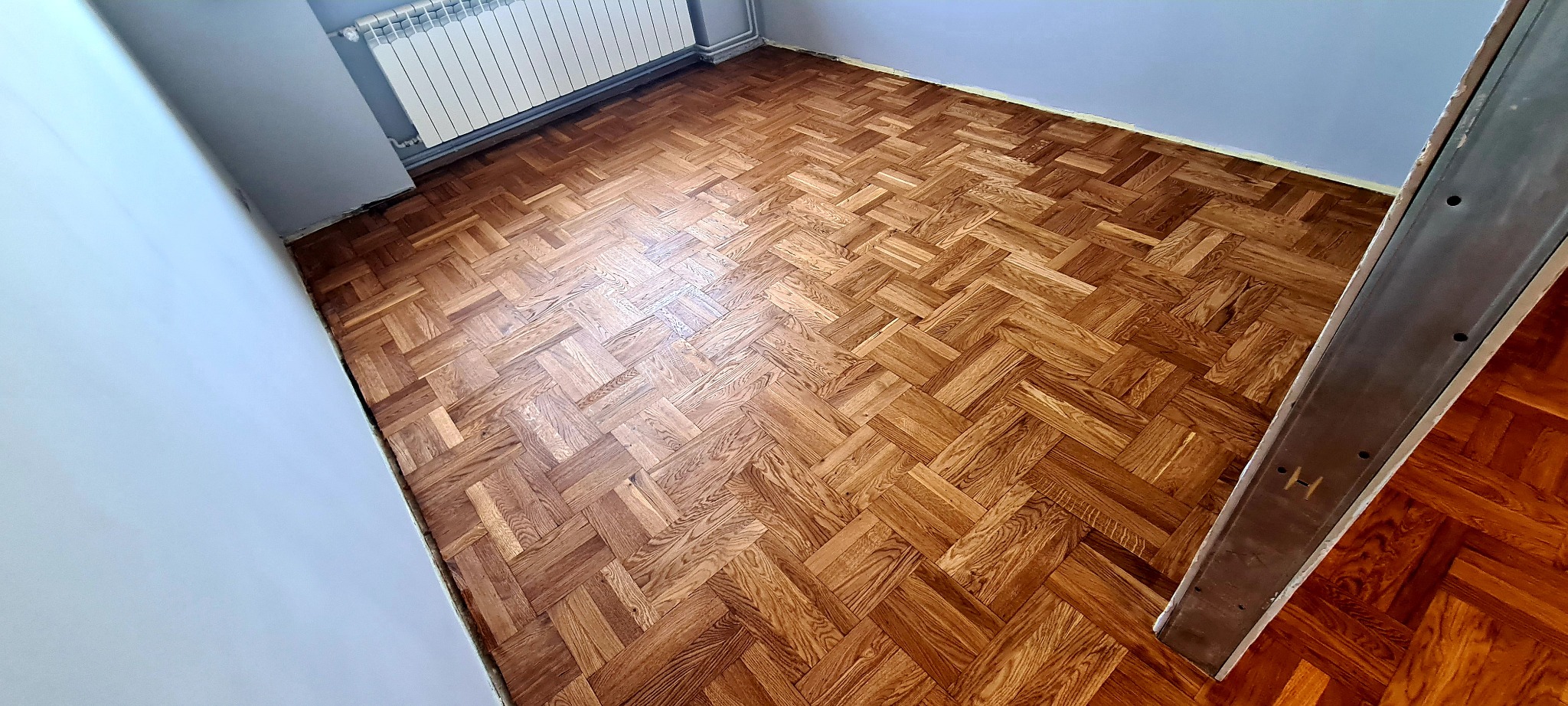 Ułożony parkiet w jodełkę z wyraźnym usłojeniem drewna, widok z góry na fragment pomieszczenia z szarymi ścianami i białym grzejnikiem.