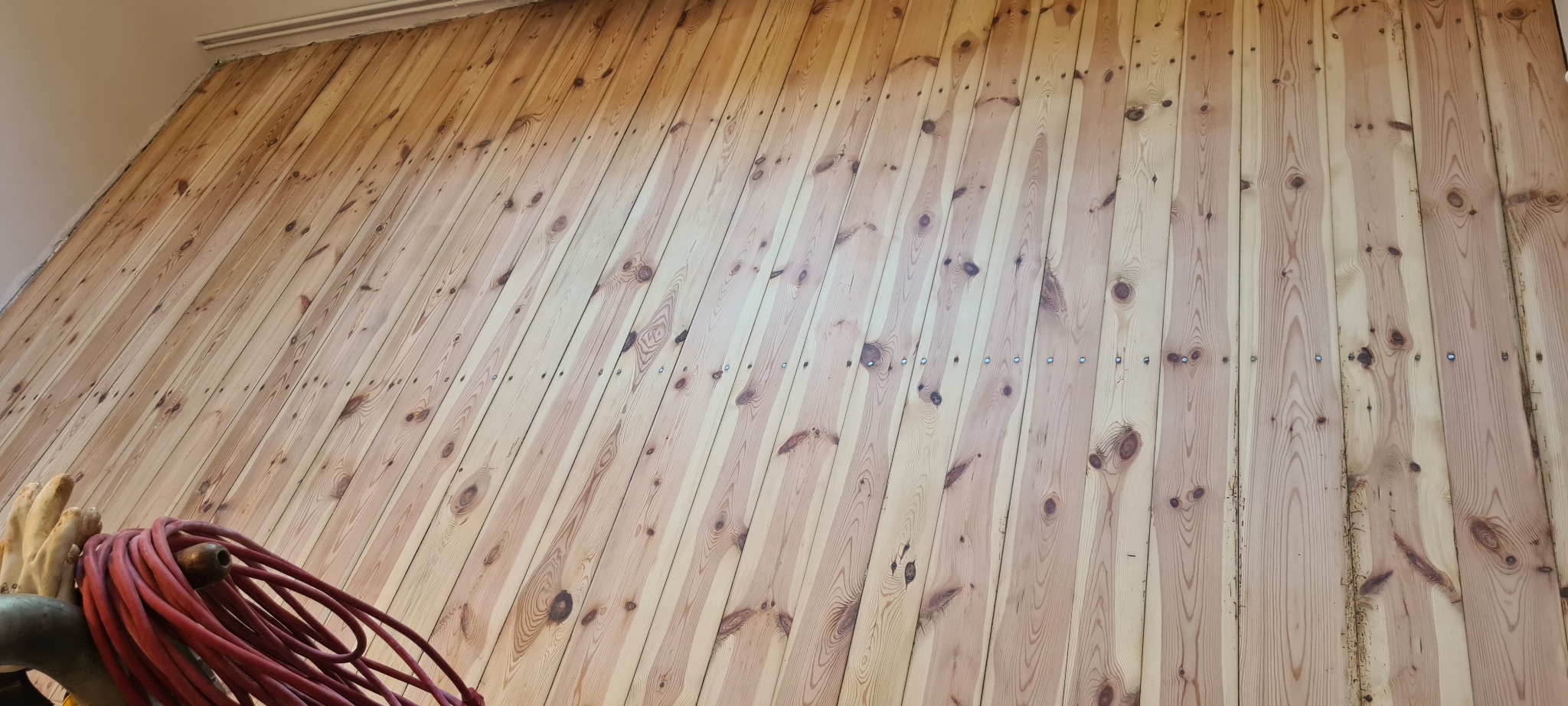 Ściana wyłożona jasnymi, pionowo ułożonymi deskami z widocznymi gwoździami, w dolnym lewym rogu widoczne narzędzia i zwinięty czerwony kabel.