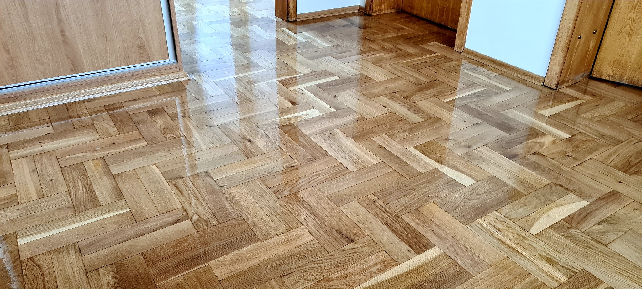 Błyszcząca, odnowiona podłoga z parkietu ułożonego w jodełkę, widoczne drzwi z jasnego drewna i fragment białej ściany.