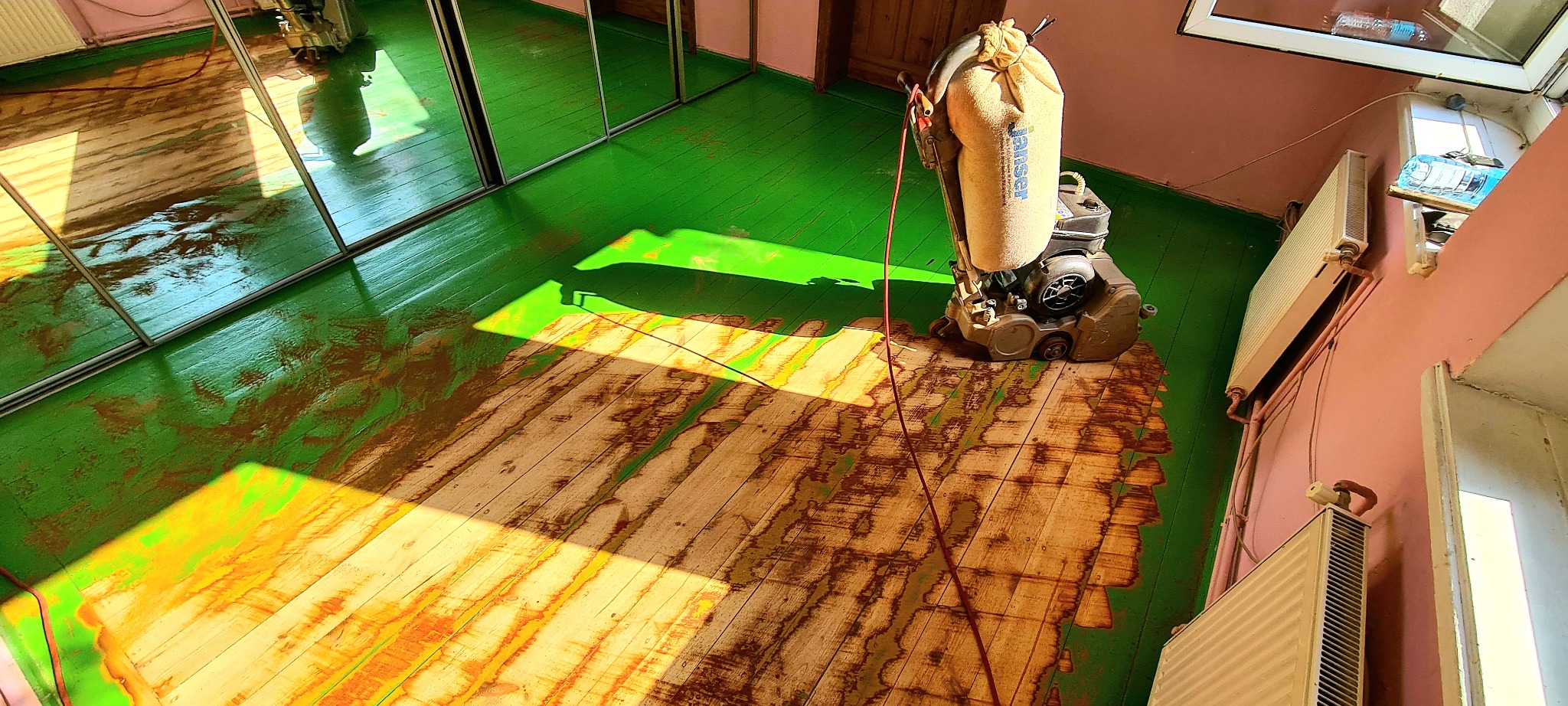 Podłoga drewniana w trakcie renowacji, częściowo wyszlifowana z zielonej farby, widoczna maszyna do szlifowania parkietu marki JanSer, jasne światło słoneczne padające na podłogę, ściany w kolorze...