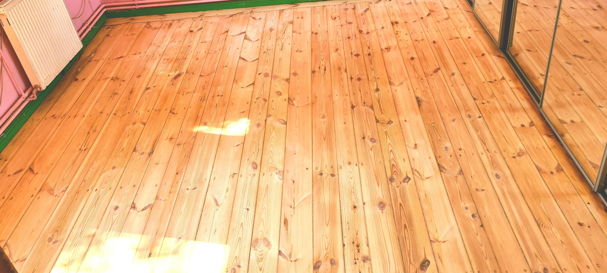 Jasna, drewniana podłoga z widocznymi słojami, po renowacji, z odbiciem w lustrze szafy przesuwnej i promieniami słońca padającymi na deski.