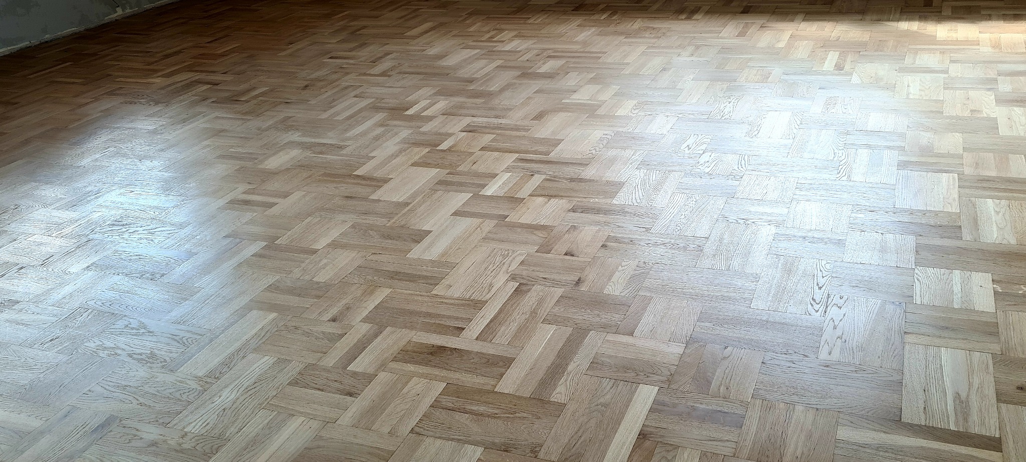 Ułożony parkiet z jasnego drewna w jodełkę francuską, widoczne niedoskonałości ściany w tle.