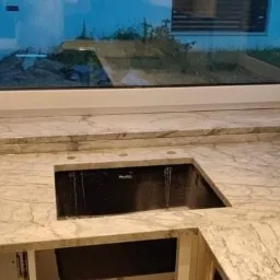 Kuchenny blat z jasnego kamienia z wyciętym otworem na zlew, zamontowany pod oknem z widokiem na zewnątrz.