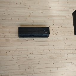 BLOWING - Czarny klimatyzator Mitsubishi Electric zamontowany na ścianie z jasnych, drewnianych paneli, widoczne dwie czarne lampy wiszące po bokach.