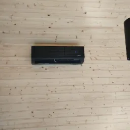 Czarny klimatyzator Mitsubishi Electric zamontowany na ścianie z jasnych, drewnianych paneli, widoczne dwie czarne lampy wiszące po bokach.