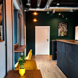 Nowoczesne wnętrze biurowe z widoczną instalacją wentylacyjną pod sufitem, oświetleniem punktowym i żółtymi akcentami kolorystycznymi w postaci krzeseł i doniczki z rośliną.