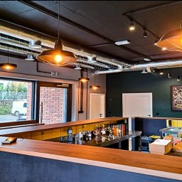 BLOWING - Wnętrze lokalu gastronomicznego z widocznymi rurami wentylacyjnymi pod sufitem, oświetleniem punktowym i wiszącymi lampami nad barem, oraz fragmentem instalacji wodno-kanalizacyjnej przy ścianie...