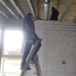 BLOWING - Montaż instalacji wentylacyjnej w surowym wnętrzu: monter na drabinie instaluje rurę wentylacyjną pod drewnianym stropem, widoczne przewody elektryczne i zielone rury wentylacyjne na podłodze.