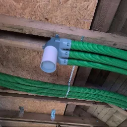 Zielone, karbowane rury wentylacyjne przymocowane do drewnianej konstrukcji stropu za pomocą metalowych uchwytów i taśmy perforowanej. Szara kształtka rozdzielająca rury z białym, cylindrycznym...